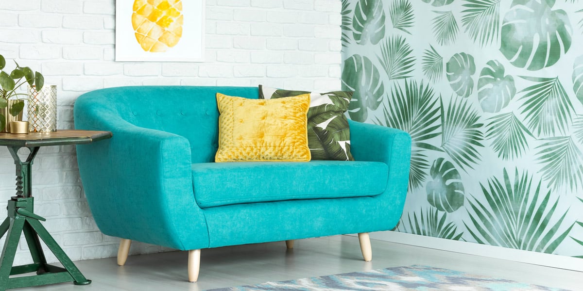 A bright teal sofa with bright, leaf-pattered throw pillows sits next to a wall treated with tropical leaf wallpaper.
